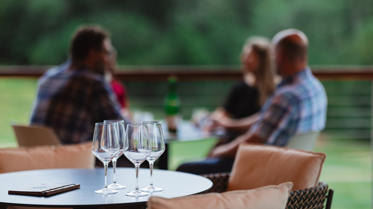 Four wine glasses in the foreground and a group of friends in the background at the Lounge.