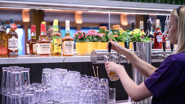 A bar tender at Meadow Lounge fills a beer glass from a tap, with glasses and liquor bottles on display.